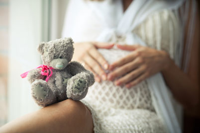 sitzende Schwangere mit ihren Händen auf dem Babybauch udn grauem Teddybär, der eine pinkfarbenes Halsband trägt, auf dem Knie